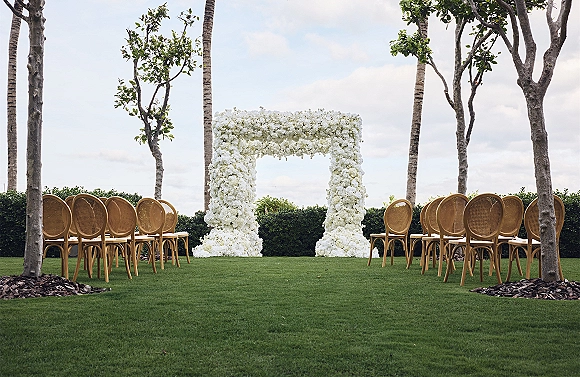 Ceremony setup with an outdoor wedding ceremony white floral arch and rattan chairs on a grass lawn with palm trees and hedges