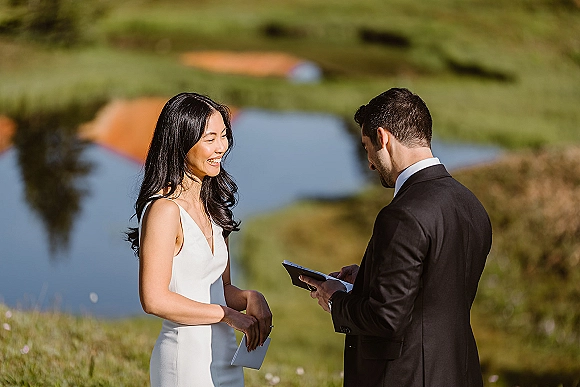 Wedding vows as bride and groom exchange paper vows, her wedding dress and his suit in a grassy field by water and hills.