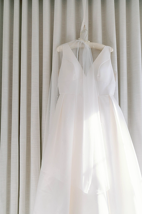 Wedding dress on hanger with bridal veil draped beside it, showcasing a plunging V neckline and full skirt against soft curtains