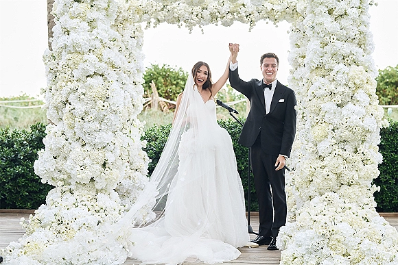 Recessional moment as newlyweds celebrating with raised hands under a white floral arch on a wooden deck, bride in veil, groom in tuxedo
