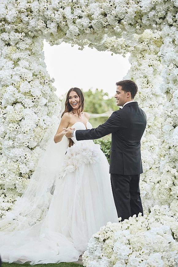 Wedding couple portrait of bride and groom holding hands, laughing beneath a white floral arch, veil flowing against bright lawn greenery