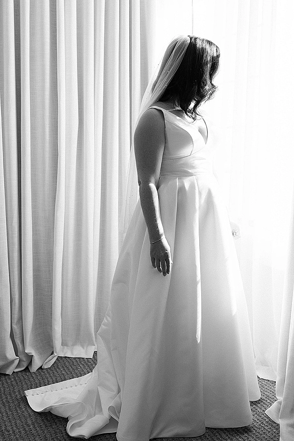 Bridal portrait in black and white of a bride by window, wearing a sleeveless satin gown and veil, bracelet visible in soft light