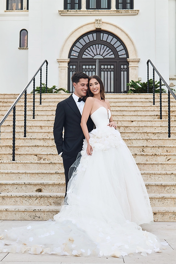 Couple portrait of bride in strapless tulle gown with long train and veil, groom in black tuxedo behind her on stone steps by arched doorway