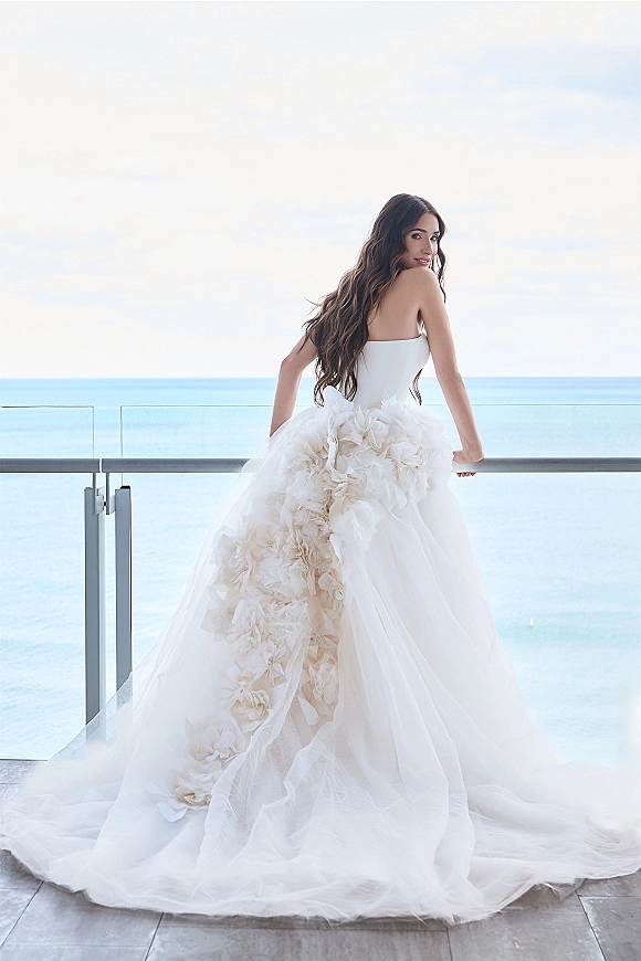Bridal portrait of a bride looking back in a strapless tulle ball gown with floral appliques on a balcony with ocean view behind her