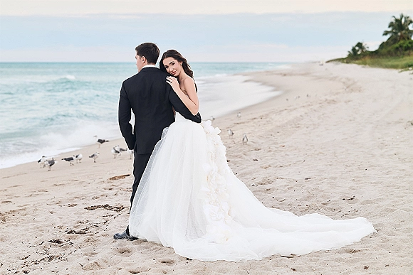 Couple portrait of bride hugging groom on a sandy beach, her veil and long dress train flowing as ocean and palm trees frame them