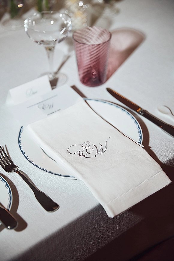 Wedding place setting with monogram wedding napkin, place card on blue-rim plate, silver flatware, wine glass and pink water glass on white linen