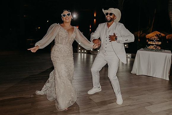 Wedding reception dancing as bride in beaded gown and heart sunglasses holds groom’s hand; he wears cowboy hat and white suit under marquee lights