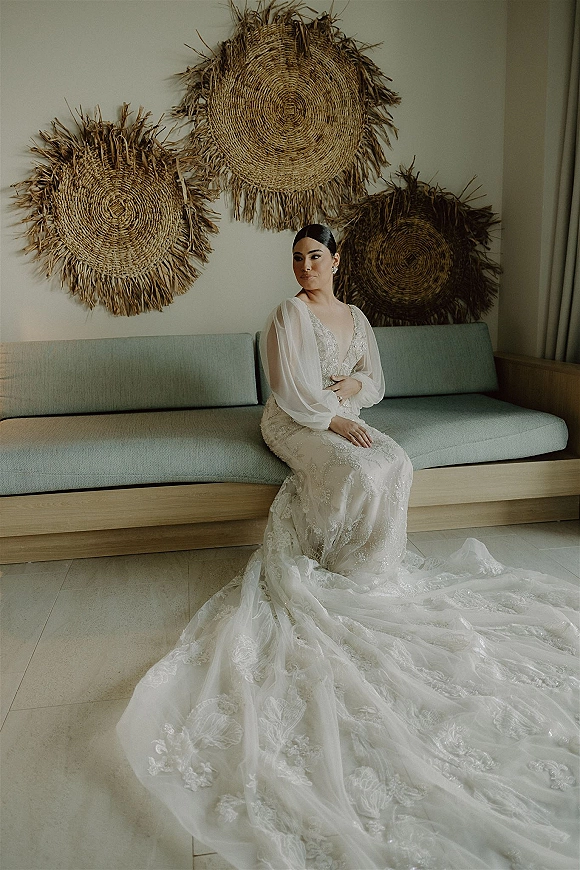 Bridal portrait of a bride sitting on sofa in a lace wedding dress with long train spread, low bun and earrings in a neutral room