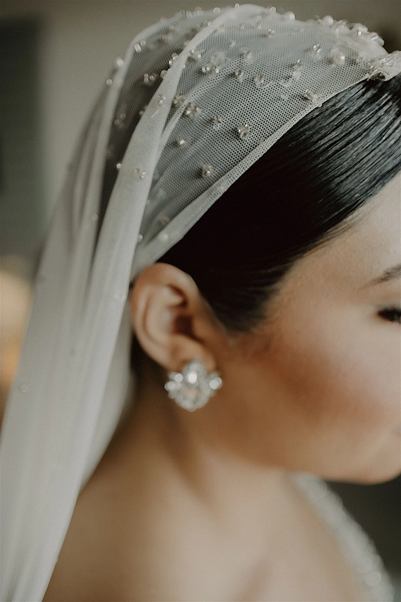 Bridal veil close-up with pearl wedding veil dots and crystal stud earrings, sheer tulle draped over an updo against a neutral wall