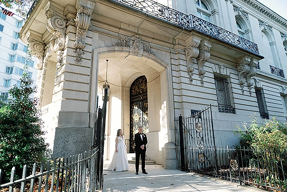 Couple portrait of bride and groom standing apart on stone steps in front of an ornate arched doorway, bride in wedding dress