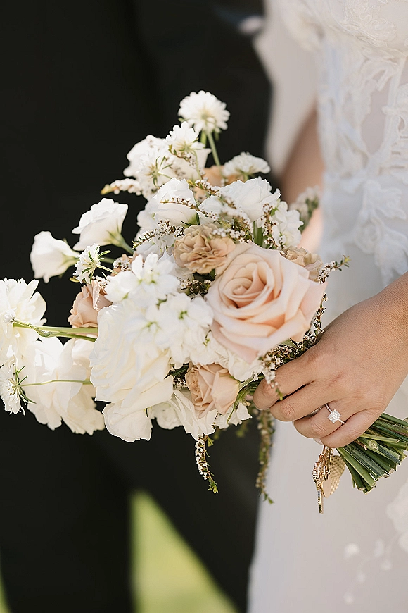Bridal bouquet of blush roses and white flowers with greenery, held against a lace dress sleeve, engagement ring, and groom suit blur