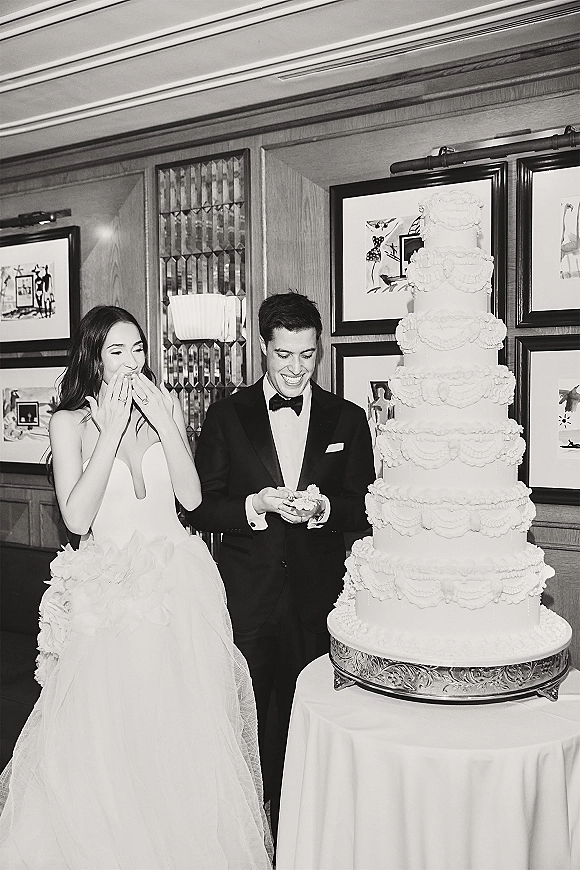 Wedding cake cutting with bride and groom cake cutting beside a tall tiered white cake on a stand, laughing in a wood-paneled reception room