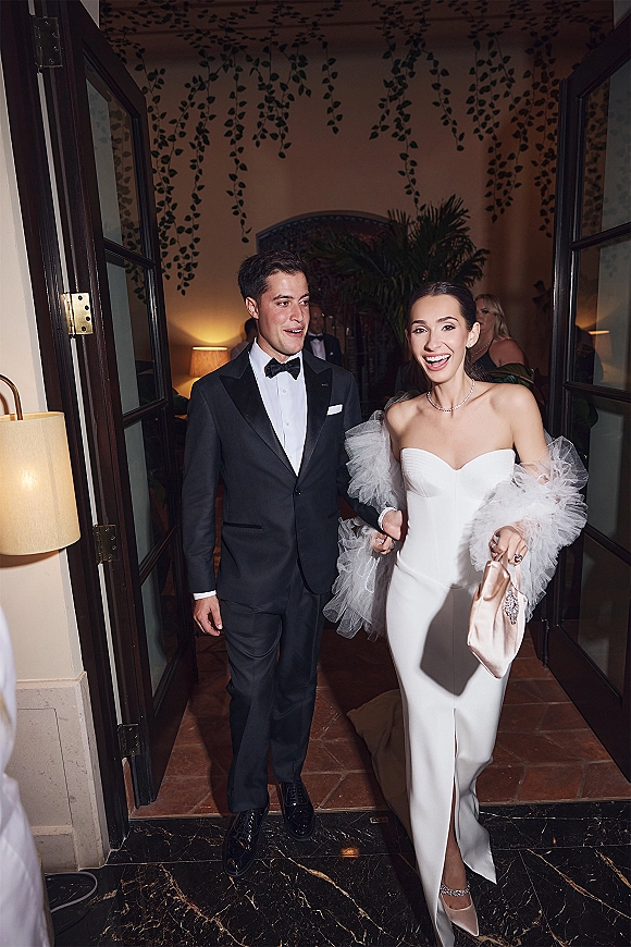 Newlywed entrance as bride laughs in a strapless wedding dress beside groom in tuxedo, walking through open doors past guests indoors