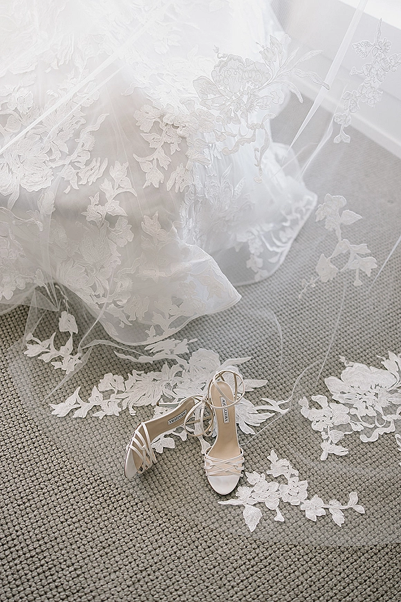 Wedding shoes in a bridal shoes flatlay with white strappy heels beside a lace wedding dress and veil on textured carpet in window light