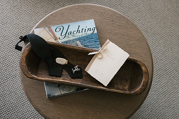 Groom accessories flat lay featuring a black necktie, silver cufflinks, tie clip, vow booklet, and wedding ring on a wooden tray table
