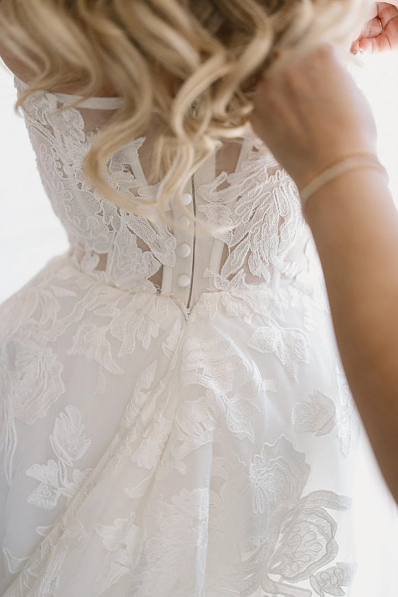 Wedding dress close-up showing lace wedding dress back with illusion panel, fabric buttons and zipper, thin straps and blonde curls in window light