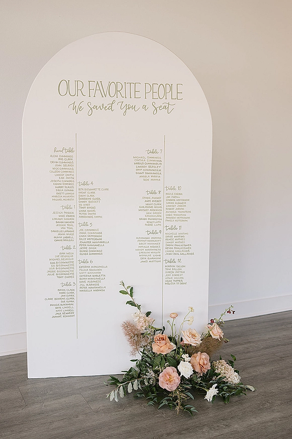 Seating chart sign in an arched seating chart style with printed guest list, blush and white flowers with greenery at base on wood floor