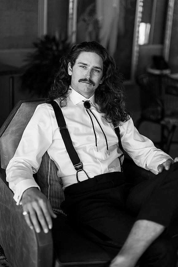 Groom portrait of a mustached, long-haired groom in suspenders and a bolo tie seated in an armchair by window light in a cozy room