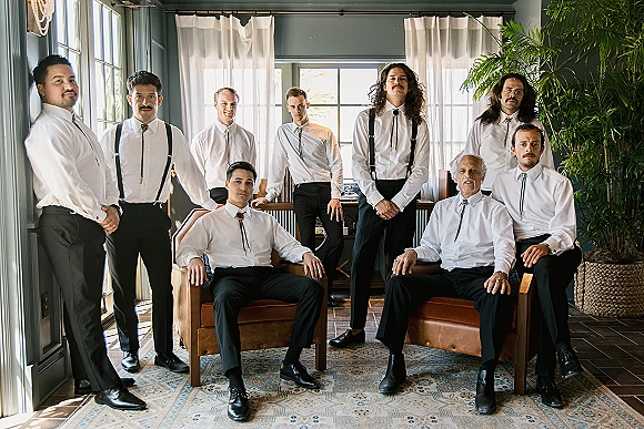 Groomsmen portrait of men in white dress shirts with bolo ties and suspenders, posed on leather chairs in a bright lounge by large windows