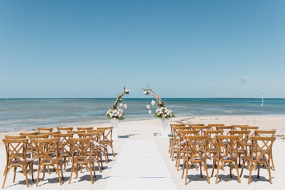 Beach ceremony setup with a white aisle runner leading to a driftwood arch, chairs on sand with ocean and blue sky beyond