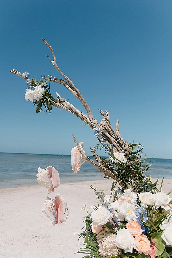 Wedding ceremony arch on a sandy beach with driftwood, white and peach roses, blue delphinium, greenery, and hanging seashells by the ocean horizon