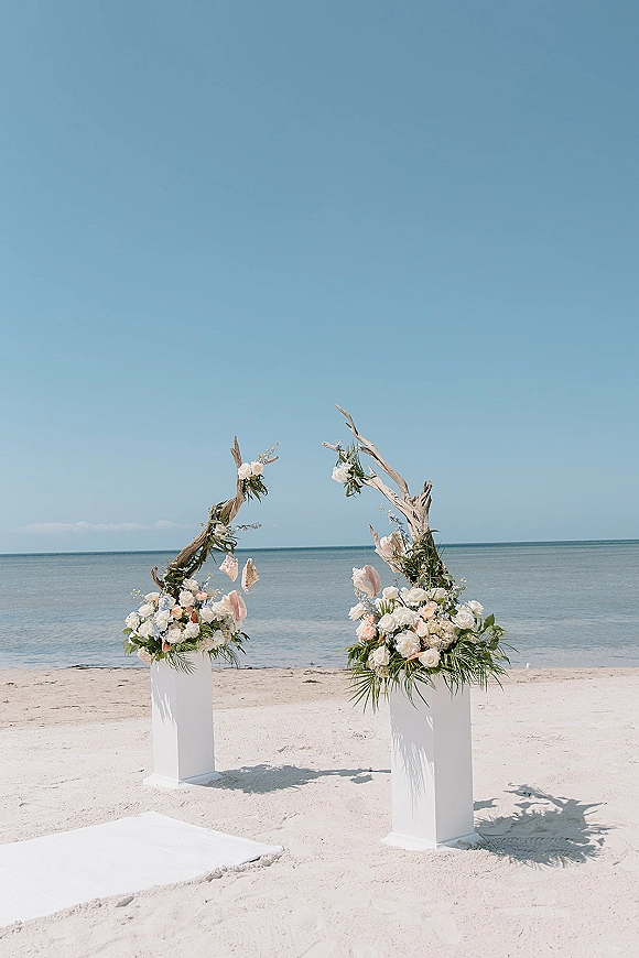 Beach wedding altar with oceanfront ceremony decor, driftwood branches, floral plinths, and a white aisle runner on sandy shore by the horizon