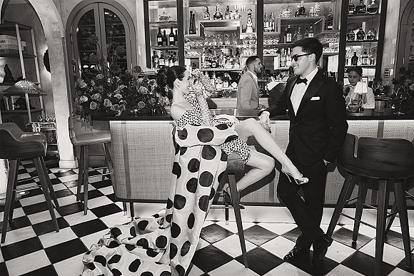 Couple portrait in a black and white wedding photo, bride in polka dot gown and gloves on bar stool beside groom in tuxedo at cocktail bar.