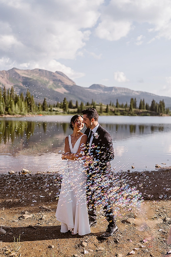 Couple portrait of bride in a sleek wedding dress and groom in a black suit smiling amid bubbles by a mountain lake with pines