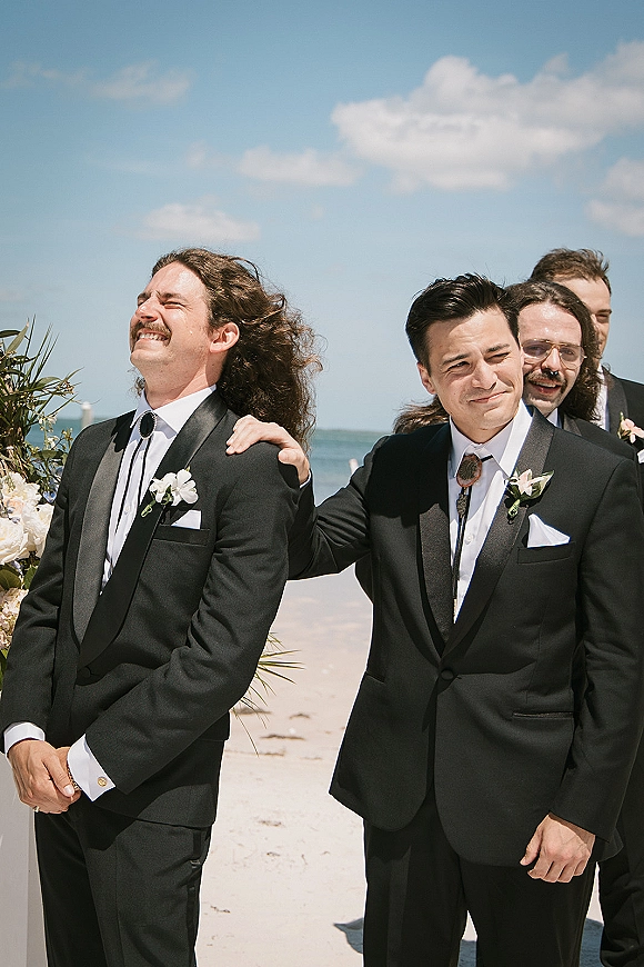 Groomsmen portrait on a beach, a groomsmen lineup photo of men in black tuxedos with boutonnières, laughing under the ocean sky