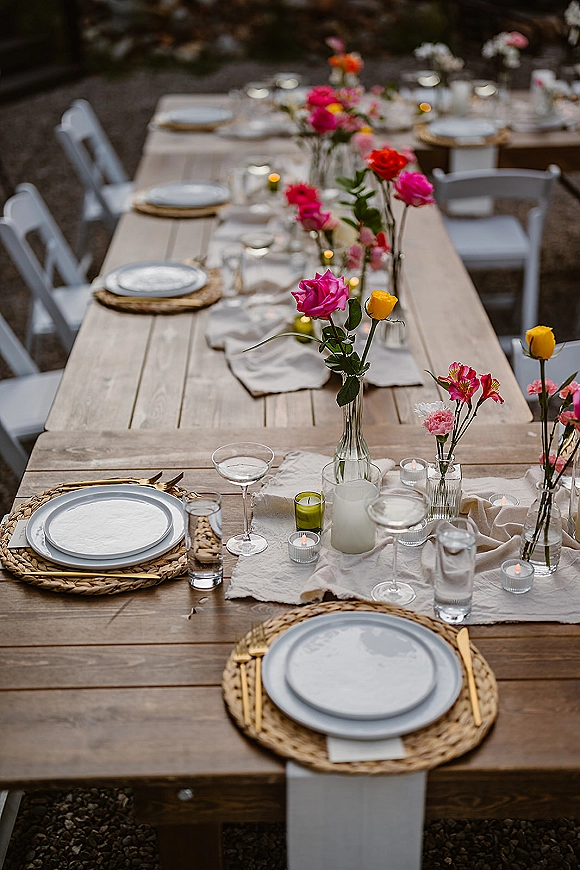 Reception tablescape on a farmhouse wedding table with rattan chargers, gold flatware, pink and yellow bud vases, and candlelight outdoors