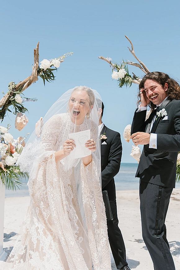Ceremony moment at a beach wedding ceremony as bride reads vows in lace dress and veil beside groom in tux under driftwood floral arch