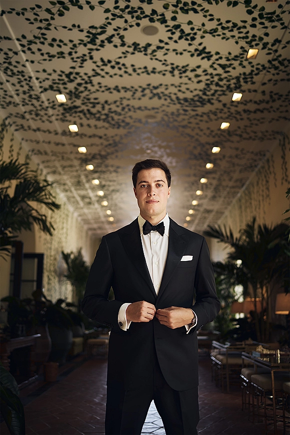 Groom portrait in a black tuxedo and bow tie, standing in an indoor hallway with patterned ceiling, lights, and potted plants