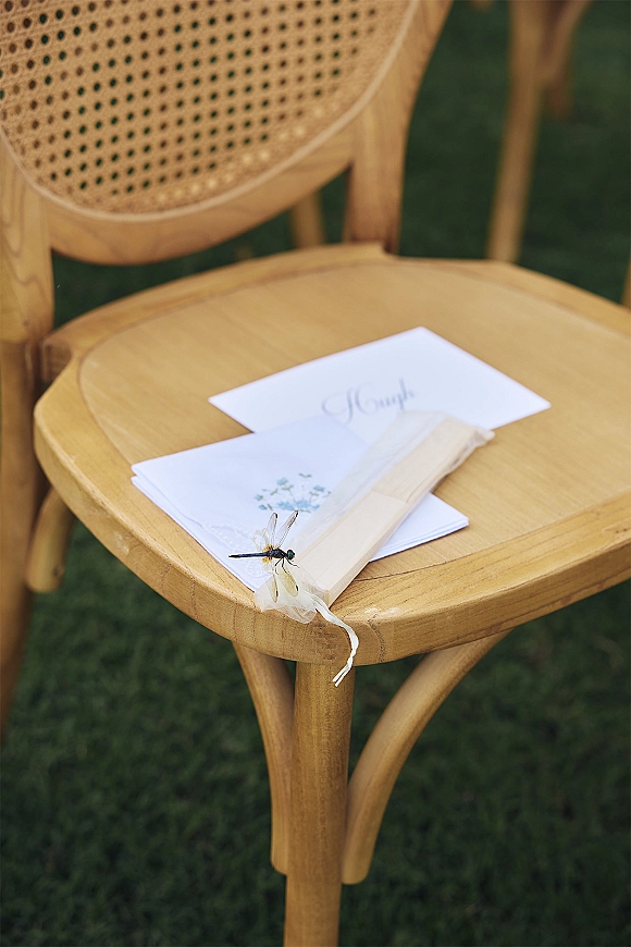 Wedding ceremony program tied with ribbon on a wooden chair with wood fan, set against outdoor seating on a grass lawn