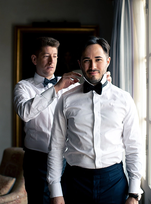 Groom getting ready as groomsmen adjust his black bow tie over a white dress shirt in window light, wristwatch visible beside curtains