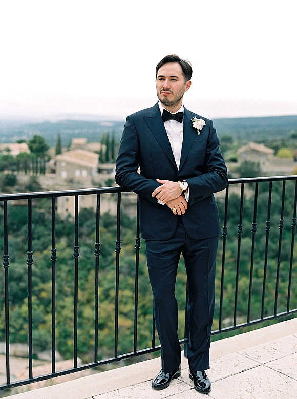 Groom portrait in tuxedo leaning on a balcony railing, wearing a black tuxedo with bow tie and boutonniere against hills and sky