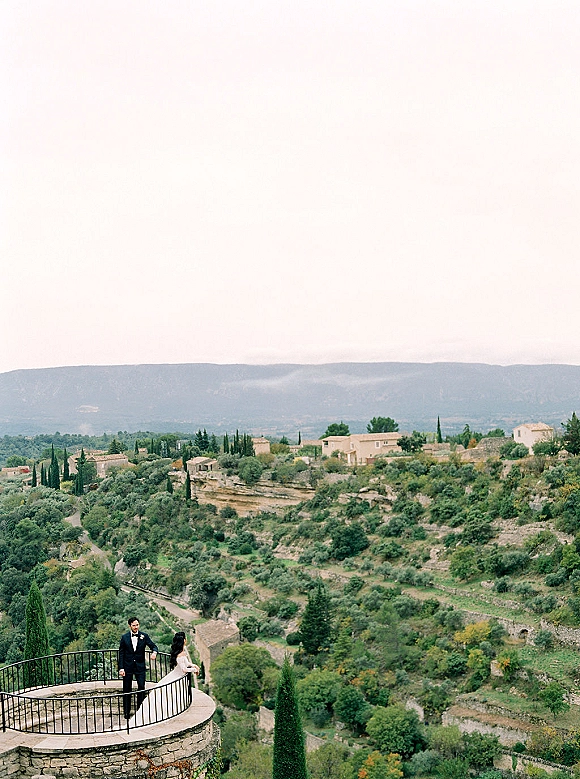 Couple portrait of bride in wedding gown and long veil with groom in tuxedo on a stone terrace overlooking a hillside village valley under cloudy sky