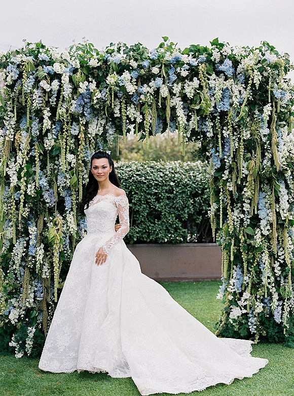 Bridal portrait of a bride in a lace wedding dress with a long train, standing under a hanging floral arch on a garden lawn