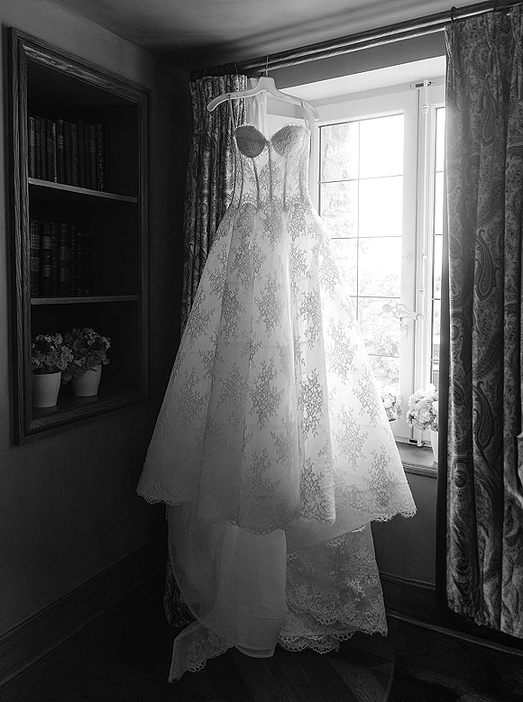 Wedding dress hanging by a window, lace wedding dress with scalloped lace accents framed by curtains, bookshelf, and flower pots