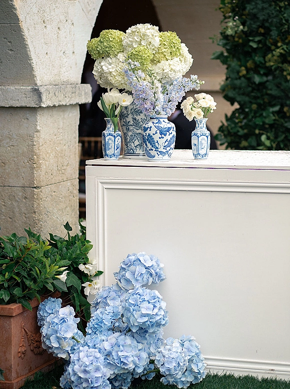 Wedding floral arrangements in blue and white chinoiserie vases with hydrangeas, delphinium and tulips on a pedestal in a stone arch courtyard