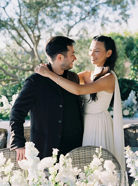 Couple portrait of bride hugging groom, both laughing in a strapless wedding dress and black suit on a garden patio with white flowers