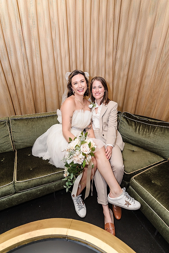 Couple portrait of a same sex wedding couple seated on a green velvet couch with bouquet, veil, beige suit, and gold curtains behind them