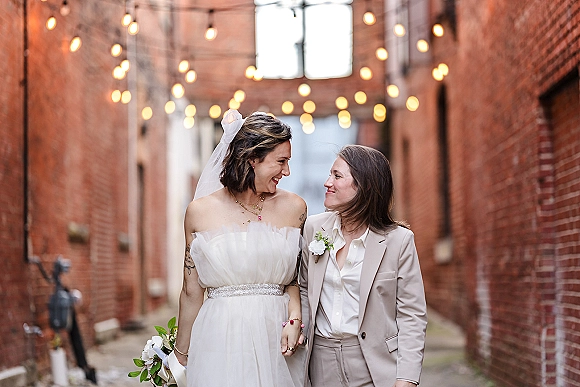 Couple portrait of a same sex wedding couple holding hands in a brick alleyway under bistro string lights, one in veil and bouquet