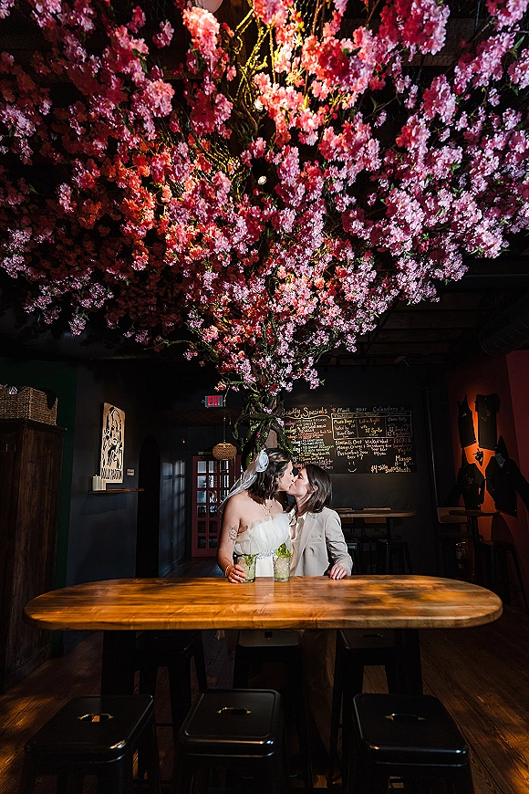 Wedding kiss portrait of two brides at a wooden table in a moody bar, veil and suit jacket, under hanging pink blossoms
