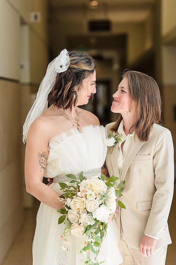 Wedding couple portrait of two brides touching foreheads, one in a veil and strapless dress holding a rose bouquet in a softly lit hallway
