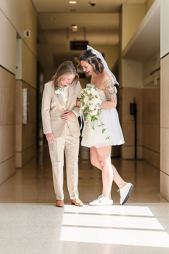 Couple portrait of a same sex wedding couple laughing as one bride adjusts a beige suit in a sunlit indoor hallway