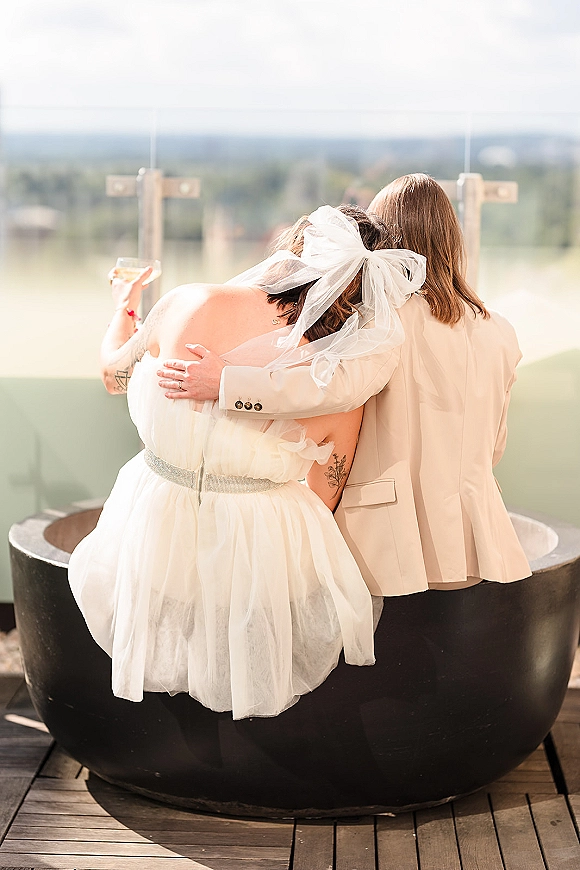 Couple portrait from behind, newlyweds sitting together on an outdoor terrace, bride in tulle bow veil and groom in white suit holding cocktails