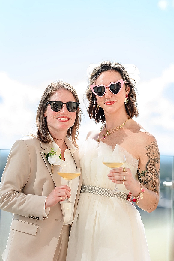 Couple portrait of two brides in veil and heart sunglasses, holding champagne coupes on an outdoor terrace with blue sky and glass railing