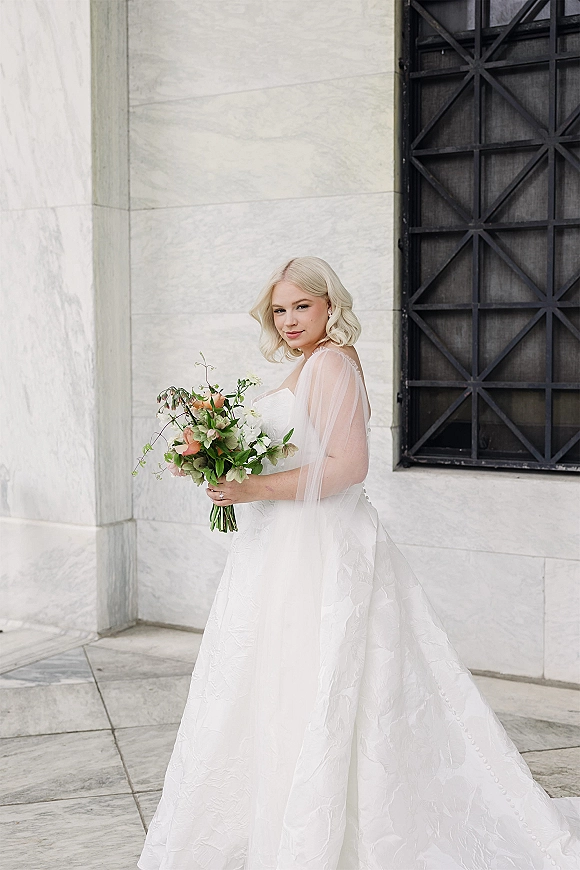 Bridal portrait of a bride holding bouquet, side profile with strapless dress and tulle veil against a stone wall and window grille