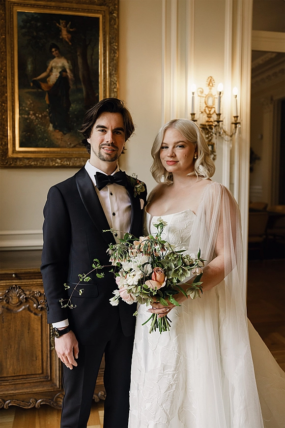 Couple portrait of bride in satin gown with cape veil and bouquet beside groom in black tuxedo, posed by framed oil painting indoors