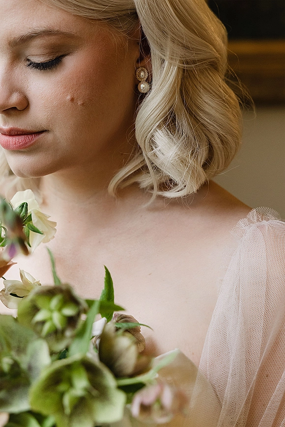 Bridal portrait of bride holding bouquet with pearl drop earrings, blonde updo, and tulle-strap gown against an indoor backdrop